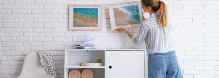 Dekorator hängt Bild auf weißem Ziegelstein Wand im Zimmer. Inneneinrichtung, weiße Kommode und weißer Stuhl, blonde Frau in Jeans hängt Bild auf